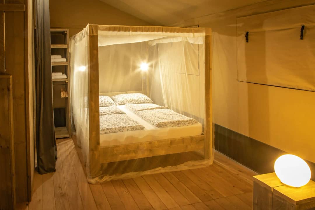 Slaapkamer van de Wood Lodge voor 2 tot 6 personen met hemelbed onder klamboe, open kast met handdoeken en een lichtbol op het nachtkastje