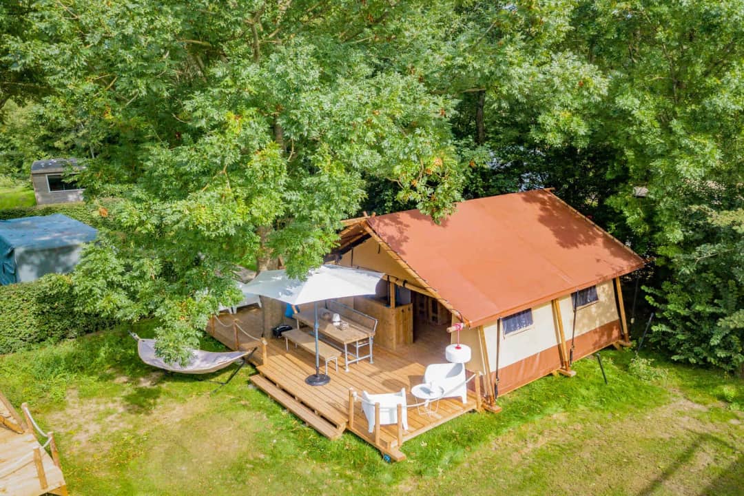 Luchtfoto van de Cozy Lodge met roodbruin dak onder hoge bomen, met parasol, eettafel en hangmat bij de veranda