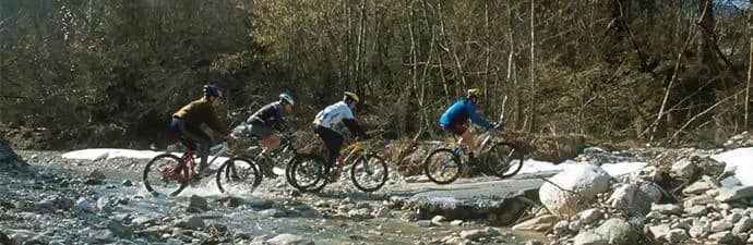 Vier mountainbikers rijden door een ondiepe beek met keien, langs kale bomen in de Veneto