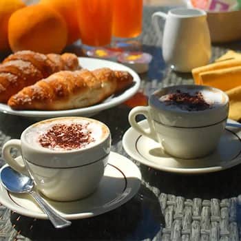 Twee cappuccino's met cacaopoeder naast croissants, sinaasappels en glazen sinaasappelsap op een ontbijttafel