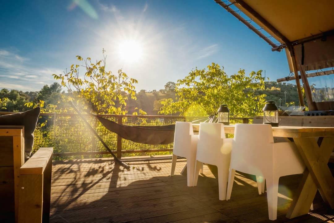 Houten eettafel met kruispoten en witte stoelen op het terras van de Desert Lodge XL, met de zon boven de bomen