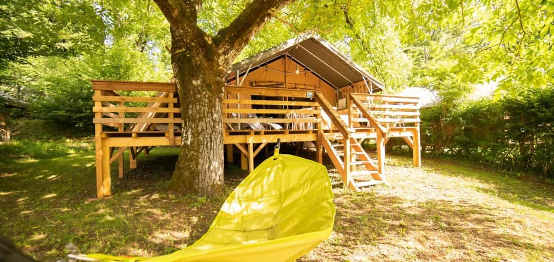 Safari Lodge op palen onder een grote boom met houten trap naar het terras en een gele hangmat op de voorgrond