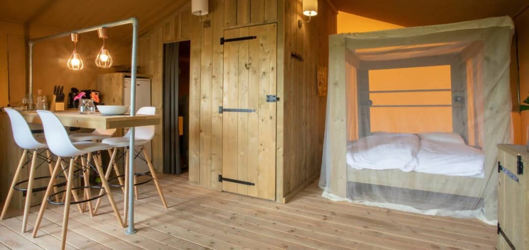 Woonruimte van de Desert Lodge XL met houten bar, witte barkrukken, houten deur en hemelbed met klamboe