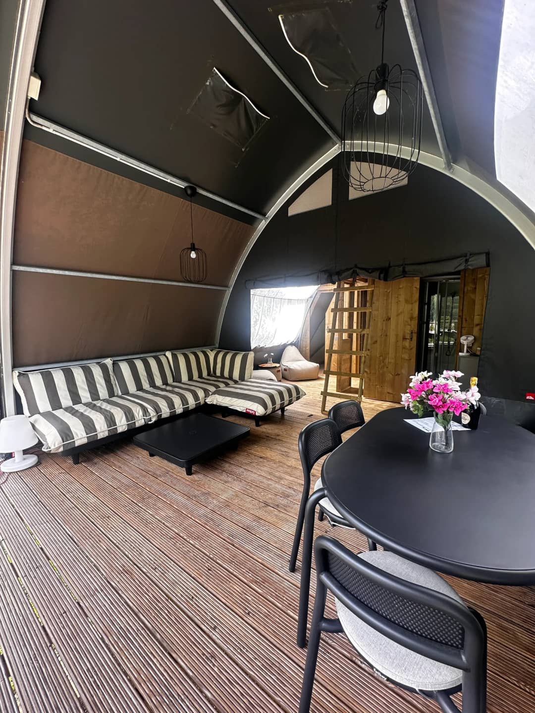 Woonruimte van de Big Oak Lodge XL met gestreepte loungebank, zwarte eettafel met bloemen en een houten ladder