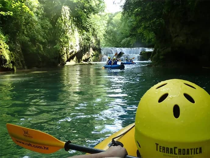 Kanotocht door een groene kloof naar een waterval, met een gele helm met de tekst TerraCroatica op de voorgrond