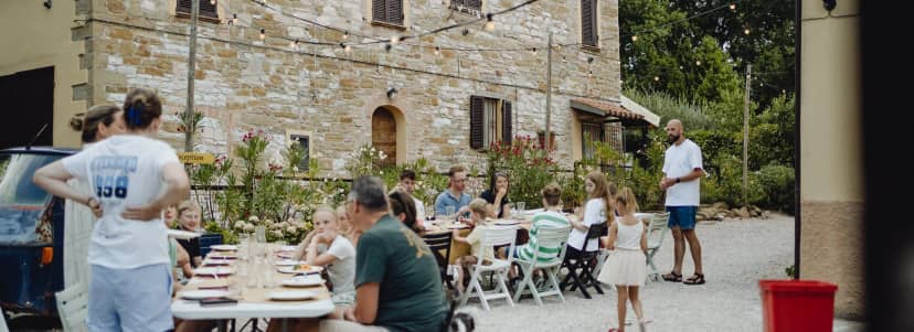Gasten eten samen aan lange tafels op het grindplein voor de stenen boerderij van Camping Casa Tartufo, onder lichtsnoeren