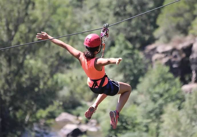 Meisje met rode helm hangt met gespreide armen aan een tokkelbaan boven de bomen