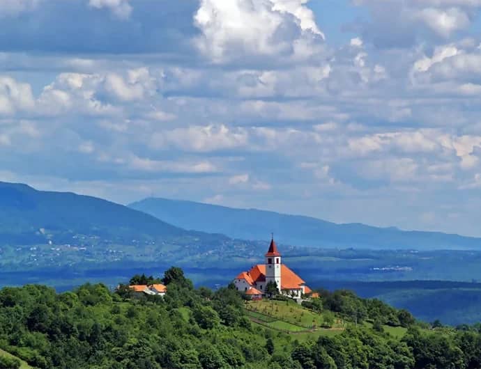 Kerk met rode torenspits op een groene heuvel, met bergen en een wolkenlucht in het binnenland van Kroatië
